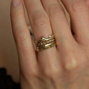 May include: A stack of four gold rings worn on a finger. The top ring features a crown-like design with small spheres. The second ring has a series of small, round, embedded stones. The third ring has a hammered texture. The bottom ring is flat.