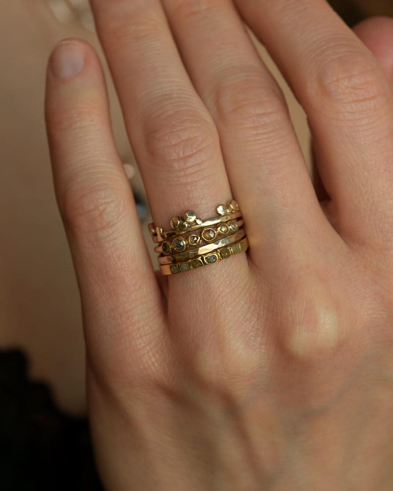 May include: A stack of four gold rings worn on a finger. The top ring features a crown-like design with small spheres. The second ring has a series of small, round, embedded stones. The third ring has a hammered texture. The bottom ring is flat.