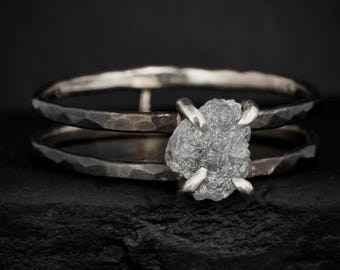 May include: A dark, oxidized silver ring featuring a double-band design with a hammered texture. The ring is set with a single, irregular, raw grey stone held in place by four prongs that secure the center piece to both bands of the ring.