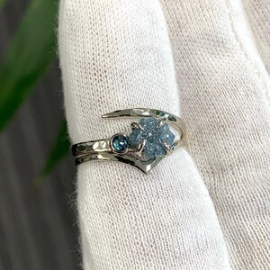 May include: A silver-toned bypass ring featuring a textured, hammered band. The ring is set with a small, round, dark blue faceted gemstone and a larger, rough-cut, light blue crystalline stone held in place by four metal prongs.