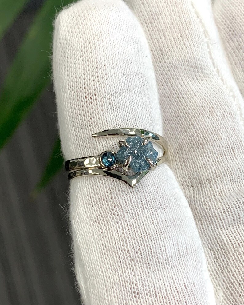 May include: A silver-toned bypass ring featuring a textured, hammered band. The ring is set with a small, round, dark blue faceted gemstone and a larger, rough-cut, light blue crystalline stone held in place by four metal prongs.