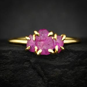 Puede incluir: Un anillo de oro con tres rubíes en bruto de color rosa vibrante dispuestos en fila horizontal. Las piedras están sujetas por pequeñas garras de oro pulido que aseguran cada pieza individual del grupo de gemas texturizadas al aro.
