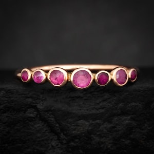 May include: A gold ring featuring seven round, bezel-set rubies of varying sizes arranged in a horizontal row. The central ruby is the largest, with three smaller rubies on each side, all set in a polished gold band with a warm, metallic finish.