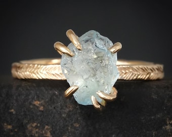 May include: A gold ring featuring a raw, light blue aquamarine stone held by four prongs. The band of the ring has a textured, repeating chevron or herringbone pattern engraved along its entire outer surface, creating a detailed metallic finish.