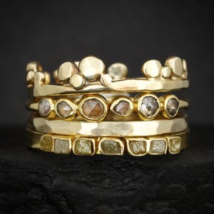 May include: A stack of four gold rings. The top ring features a series of smooth, rounded gold spheres. The second ring contains a row of six bezel-set, faceted brown and grey stones. The third ring is a plain, hammered gold band. The bottom ring holds six square-cut, pale yellow raw stones.
