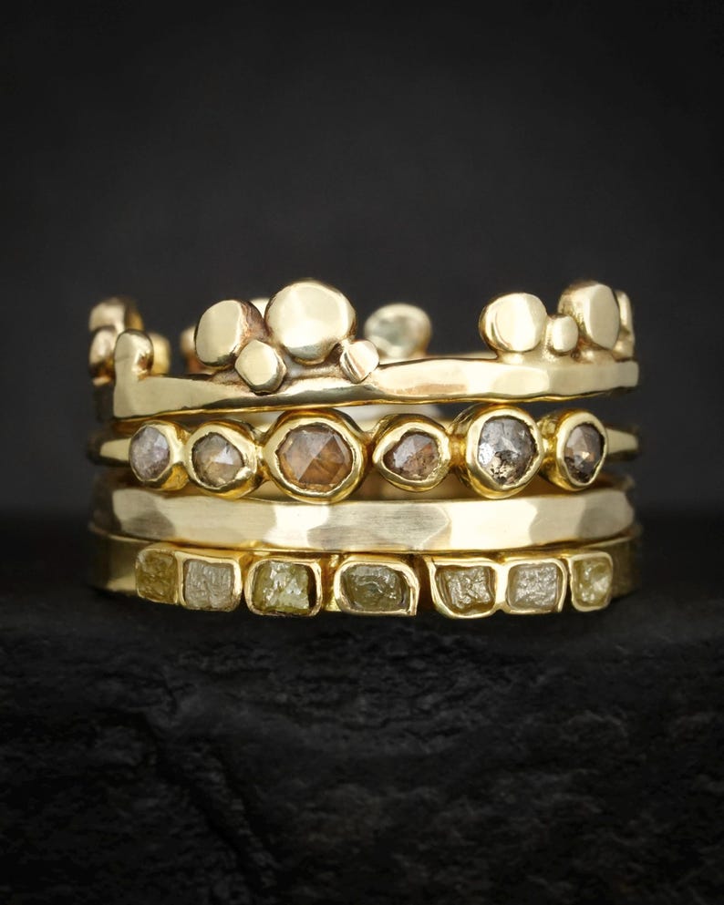 May include: A stack of four gold rings. The top ring features a series of smooth, rounded gold spheres. The second ring contains a row of six bezel-set, faceted brown and grey stones. The third ring is a plain, hammered gold band. The bottom ring holds six square-cut, pale yellow raw stones.