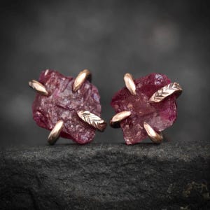 Peut inclure: Une paire de boucles d'oreilles à clous avec des pierres précieuses brutes rouge rosé foncé maintenues par des griffes métalliques. Les griffes présentent un motif texturé en forme de feuille. Les pierres ont une forme irrégulière et dentelée.