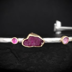 May include: A silver cuff bracelet with a hammered texture, featuring a central raw, deep pink gemstone set in gold, flanked by two smaller, round, faceted pink gemstones also set in gold bezels along the band.