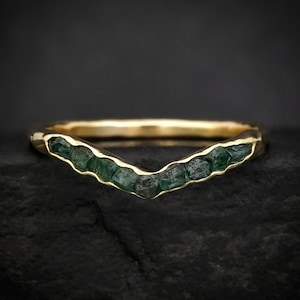 May include: A gold ring with a V-shaped design. The ring features a row of dark green gemstones. The ring is presented on a dark, textured surface.