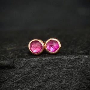 Könnte beinhalten: Ein Paar runde Ohrstecker mit leuchtend pinken, facettierten Edelsteinen, die in polierte Fassungen aus Roségold eingefasst sind. Die kreisförmigen Steine zeigen einen tiefen Magentaton mit sichtbaren inneren Facetten, die das Licht reflektieren.