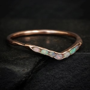 May include: A V-shaped ring crafted from rose-toned metal, featuring a series of small, iridescent opals embedded along the front curve. The band has a smooth, polished finish, and the stones display subtle flashes of green, blue, and pink light.