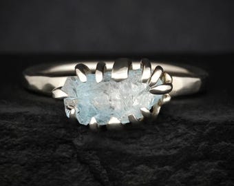 May include: A ring featuring a raw, light blue aquamarine gemstone held in place by multiple silver-toned metal prongs. The band of the ring has a smooth, polished finish and a rounded profile, showcasing the natural texture of the stone.