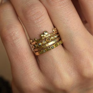 May include: A stack of four gold rings worn on a finger. The top ring features a crown-like design with small gold spheres. The middle rings include one with embedded dark stones and one with a hammered texture. The bottom ring has a row of green stones.