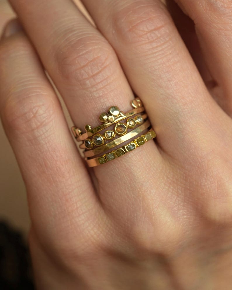May include: A stack of four gold rings worn on a finger. The top ring features a crown-like design with small gold spheres. The middle rings include one with embedded dark stones and one with a hammered texture. The bottom ring has a row of green stones.