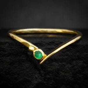 May include: A gold ring with a V-shaped design. The ring features a small, round, green gemstone set in the centre. The band has a hammered texture, and the ring is displayed against a dark background.