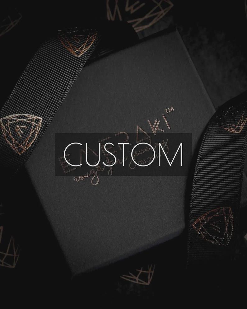 May include: A square black gift box with the word CUSTOM printed in white across the centre. The box features a black ribbon with a repeating copper-coloured geometric logo pattern. The brand name is embossed in copper on the box lid surface.