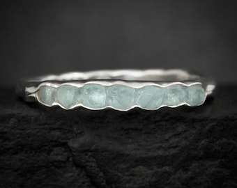 May include: A silver ring featuring a row of six small, light blue, raw, uncut gemstones set horizontally along the band. The metal band has a textured, organic appearance with a polished finish, showcasing the natural shape of the stones.
