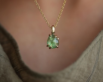 May include: A gold chain necklace featuring a faceted, pear-shaped green gemstone pendant. The stone is secured in a gold prong setting that holds the gem at the base and sides, suspended from a delicate gold link chain with a polished finish.