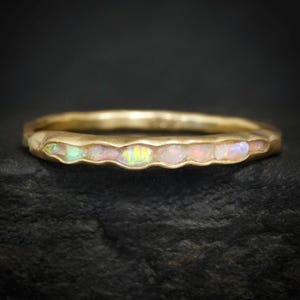 Może przedstawiać: Złoty pierścionek z teksturowanym paskiem i rzędem opalów. Opale prezentują gamę kolorów, w tym różowy, biały i odcienie zieleni. Pierścionek jest umieszczony na ciemnym tle.