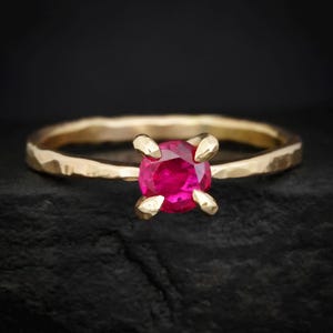 May include: A gold ring with a hammered band and a vibrant, round ruby gemstone. The ruby is held in place by four prongs, creating a classic and elegant design. The ring is set against a dark background.