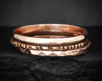 May include: A stack of three rose gold rings. The top ring has a smooth, polished surface. The middle ring features a textured, notched pattern around the band. The bottom ring has a faceted, hammered finish with multiple flat surfaces.