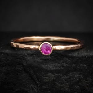 Könnte beinhalten: Ein dünner Ring in Roségold-Optik mit einem gehämmerten, strukturierten Band und einem einzelnen, kleinen, runden, leuchtend pinkfarbenen Edelstein, der in einer einfachen Zargenfassung in der Mitte des Rings sitzt und ein zartes Design aufweist.