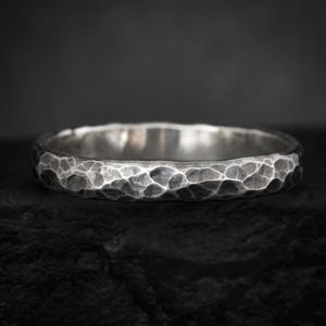 May include: A silver ring featuring a dark, oxidised finish and a textured, hammered surface. The band has a rounded profile with irregular indentations across the exterior, creating a high-contrast appearance between the bright and dark metal.