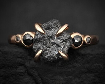May include: A rose gold ring featuring a large, central, rough-cut black diamond held by four prongs. The band includes two smaller, faceted black diamonds on either side, followed by two tiny, round black stones set into the metal band.