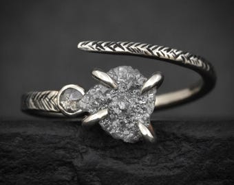 May include: A bypass ring featuring a textured, dark metal band with a herringbone pattern. The ring is set with a large, rough, grey, pear-shaped diamond held by four prongs and a smaller, round, faceted grey diamond set in a bezel.