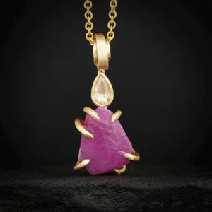 Puede incluir: Collar con colgante de oro con una gran piedra preciosa rosa de forma irregular. La piedra preciosa está acentuada con garras doradas y una piedra transparente en forma de lágrima. La cadena es de oro fino.
