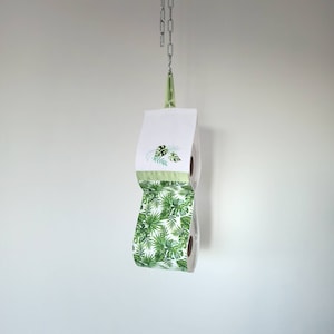 May include: A white double toilet roll holder with a green tropical leaf pattern. The holder is hanging from a silver chain.