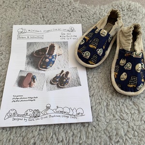 May include: A pair of blue and white children's slippers with a cafe theme. The slippers have a white trim and are made of fabric. The slippers are shown on a white sheet of paper with instructions for making them. The paper also includes the text "Children's Slippers Cuk 6C-2A", "Sole oos", "Size Eu27/21", "(fits UK 9-4.0C)", "Pattern & Instructions", and "Designed by Gerli Lau © 2017 Mushroom Village Crafts".