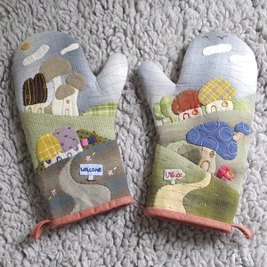 Puede incluir: Un par de manoplas de horno coloridas con un tema de hongos y casas. Las manoplas están hechas de tela de retazos y tienen las palabras "Bienvenido" y "Pueblo".