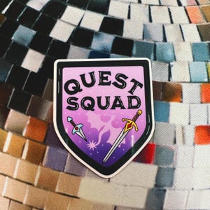 May include: A pink and purple shield-shaped sticker with the text "QUEST SQUAD" in black lettering. Two swords, one blue and one gold, are crossed behind the text.