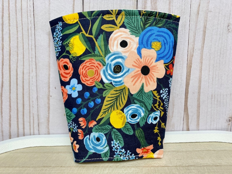 May include: A navy blue fabric cup cosy with a floral pattern featuring pink, orange, yellow, and blue flowers. The fabric has a white background with a dark blue outline.