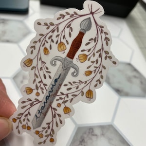 May include: A white sticker with a silver sword surrounded by a wreath of brown branches and yellow flowers. The sword has the words "Ataraxia Inner Peace" written on it.