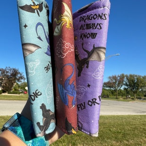 May include: Three fabric wristlets with dragon designs. The wristlets are in shades of blue, purple, and red. The purple wristlet has the text "DRAGONS ALWAYS KNOW FLY OR DIE".