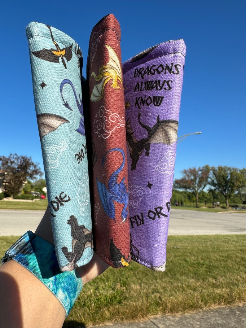 May include: Three fabric wristlets with dragon designs. The wristlets are in shades of blue, purple, and red. The purple wristlet has the text "DRAGONS ALWAYS KNOW FLY OR DIE".