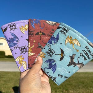May include: Three fabric can koozies with a dragon theme. The koozies are purple, red, and blue with a white background. The text on the koozies reads "Dragons Always Know" and "Fly or Die".
