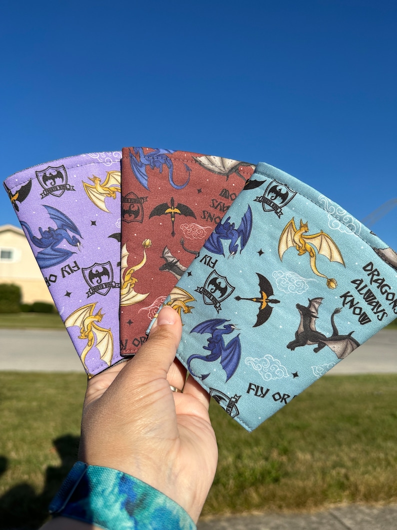 May include: Three fabric can koozies with a dragon theme. The koozies are purple, red, and blue with a white background. The text on the koozies reads "Dragons Always Know" and "Fly or Die".