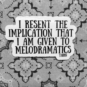Puede incluir: Una pegatina blanca con texto negro: "I RESENT THE IMPLICATION THAT I AM GIVEN TO MELODRAMATICS", con "TAIRN" debajo. La pegatina est&aacute; sobre un fondo con estampado gris y blanco.