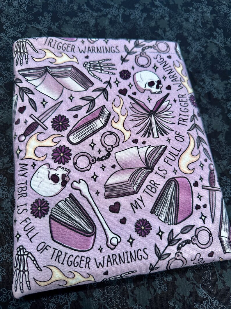 Pu&ograve; includere: Tessuto viola con un motivo ripetitivo di libri, teschi, ossa, manette, fiamme e fiori. Il testo "My TBR is full of trigger warnings" &egrave; ripetuto in tutto il motivo.