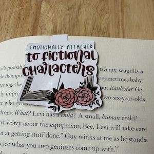 May include: White bookmark with black and pink floral design. The bookmark has the text "Emotionally attached to fictional characters" in brown lettering.