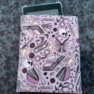 Pu&ograve; includere: Una custodia per libri rosa e bianca con un motivo di libri, teschi, cuori e fiamme. Il testo sulla custodia recita "My TBR is full of trigger warnings".