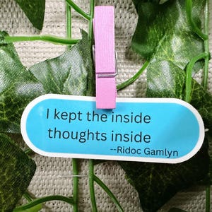 May include: A blue and white oval sticker with the quote "I kept the inside thoughts inside" attributed to Riddoc Gamlyn. The sticker is attached to a green leafy background with a pink clothespin.