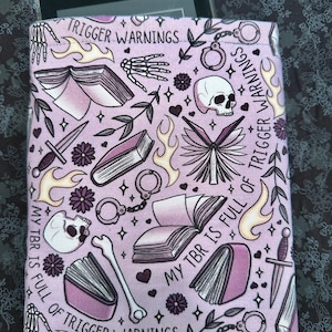 May include: A pink fabric book sleeve with a repeating pattern of books, skulls, bones, handcuffs, flowers, and flames. The text "My TBR is full of trigger warnings" is repeated on the sleeve.