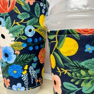 May include: Two reusable coffee cup sleeves with a floral pattern. The sleeves are made of fabric and have a navy blue background with colourful flowers, leaves, and lemons.