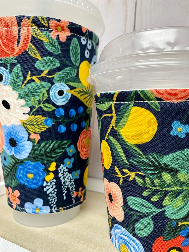May include: Two reusable coffee cup sleeves with a floral pattern. The sleeves are made of fabric and have a navy blue background with colourful flowers, leaves, and lemons.