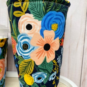 May include: A reusable coffee cup sleeve with a floral pattern. The sleeve is made of fabric and has a dark blue background with pink, blue, yellow, and green flowers.