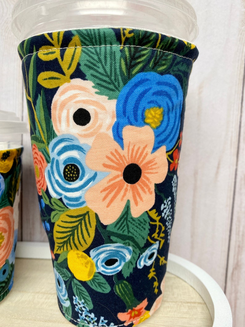 May include: A reusable coffee cup sleeve with a floral pattern. The sleeve is made of fabric and has a dark blue background with pink, blue, yellow, and green flowers.
